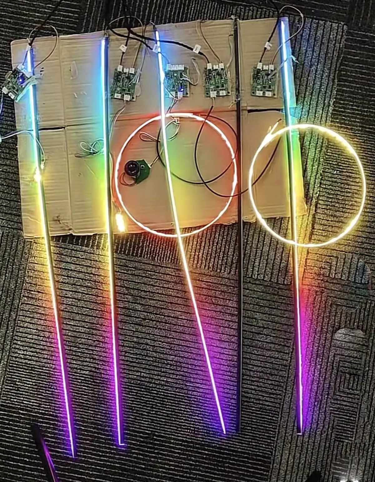 Five fibre-optic strips and two circular halo rings laid out on cardboard, all lit in a full rainbow gradient, each strip wired to its own driver board.