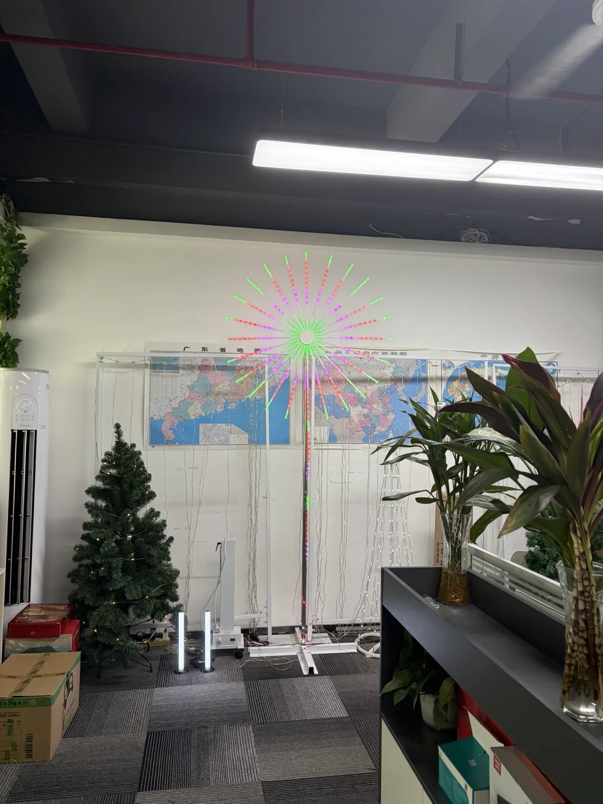 A firework LED light mounted on a pole in an office, its radiating strands lit green and pink, with a Christmas tree and other decorative lights around it.