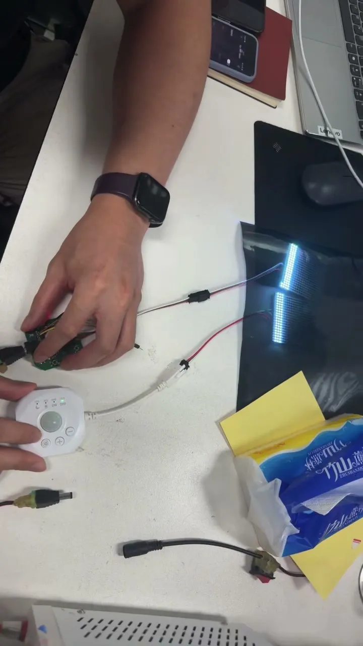 A hand holding a round controller next to a phone and a lit LED strip on a desk during pairing.