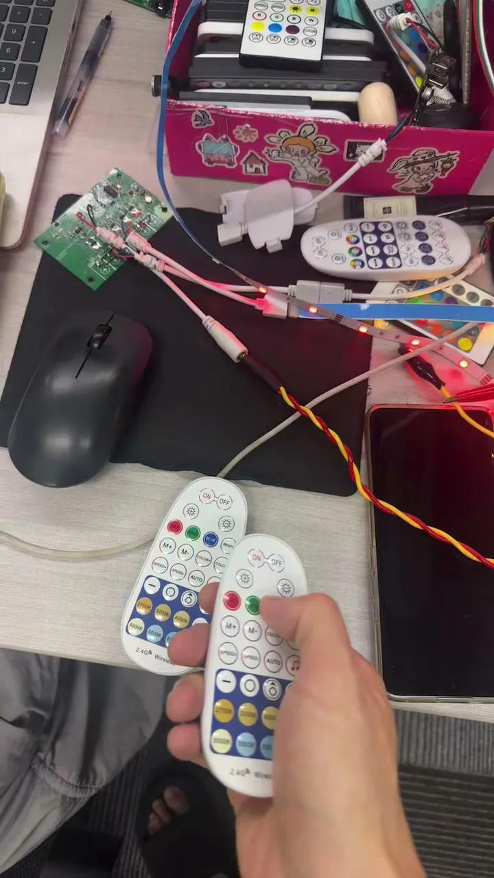 A hand holding an RF remote in front of LED strips lit blue on a desk.