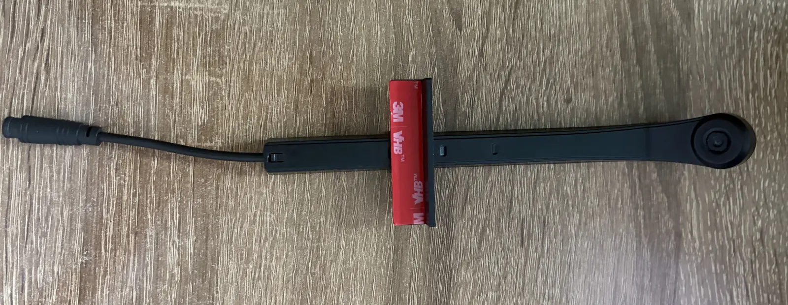 The screen colour sensor of a TV backlight kit laid on a table: a camera head on an arm, a cross bracket carrying red 3M VHB tape with its liner still on, and a DC lead.