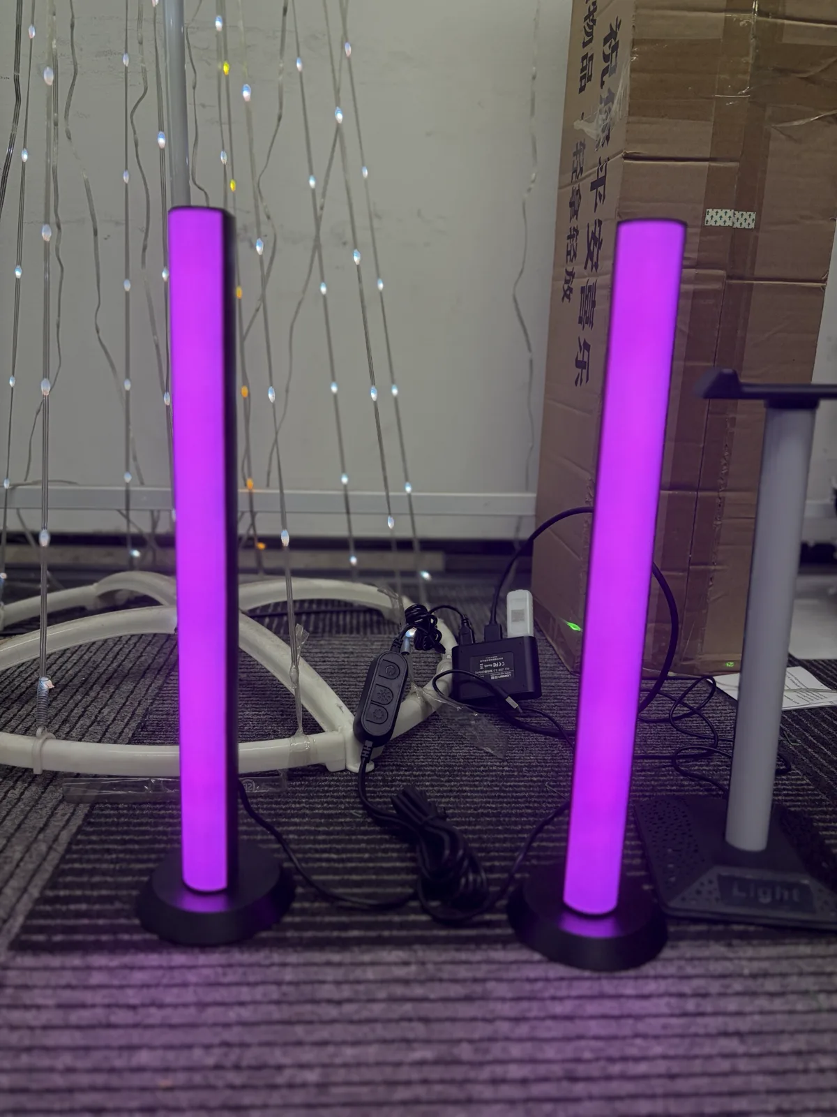 Two sound-activated LED bars standing on a carpeted floor, both lit magenta, with an inline controller and a mains power adapter on the cable between them.