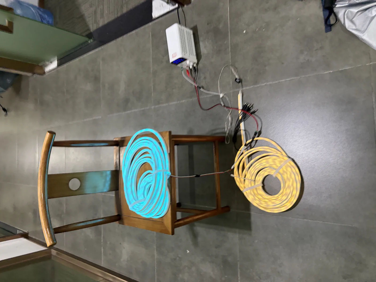 Two coils of silicone LED neon flex on test, one lit cyan and one lit warm yellow, wired back to a bench power supply showing its voltage and current readout.